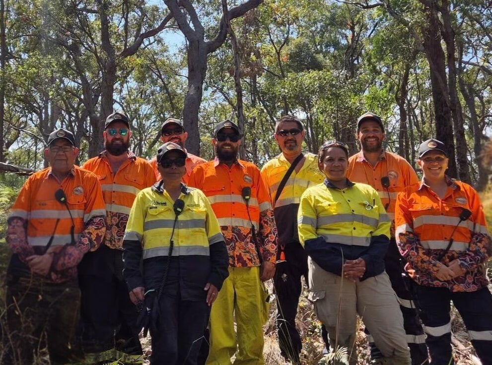 A group of nine rangers and fire management personnel in high-visibility orange and yellow uniforms posing together in a sunny, wooded area.